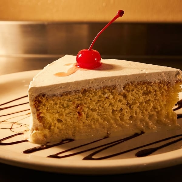 Slice of tres leches cake with white frosting, a cherry, and chocolate drizzle on a white plate.