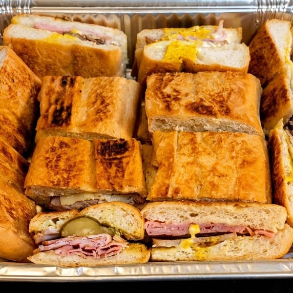 A catering tray filled with many Cuban sandwiches, cut in halves and quarters, showing ham, cheese, and pickles.