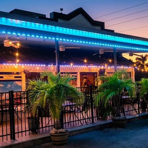Exterior of La Teanila restaurant at dusk, with blue and string lights, palm trees, and outdoor seating.