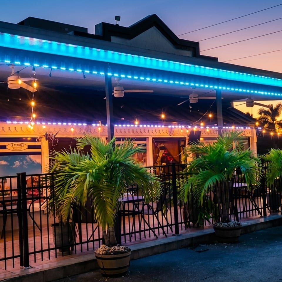 Exterior of La Teanila restaurant at dusk, with blue and string lights, palm trees, and outdoor seating.